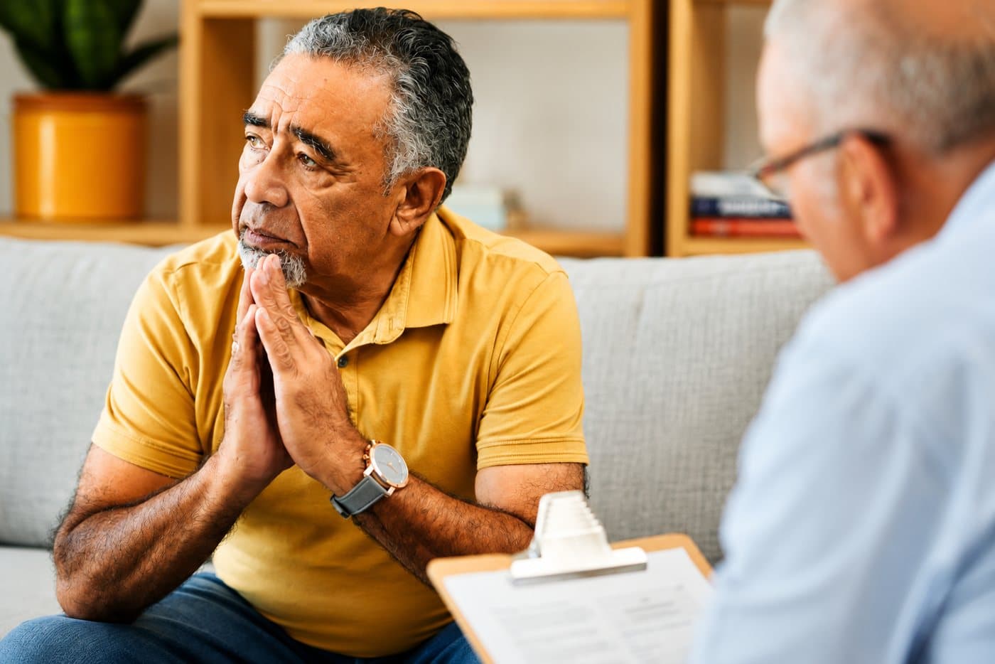 An older man sitting with his hands clasped, talking with a counselor