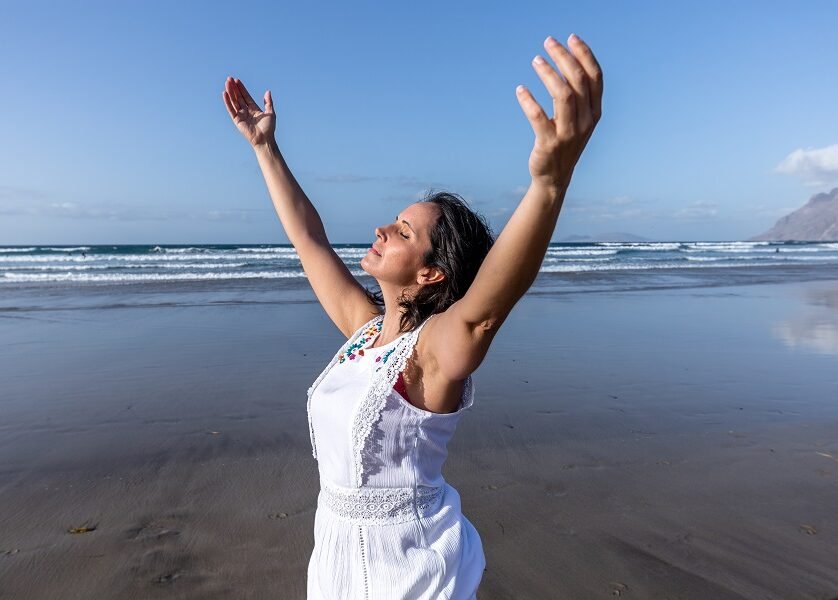 Middle aged female traveler raising arms and breathing fresh air with closed eyes while resting on wet beach near waving sea on sunny summer day on resort