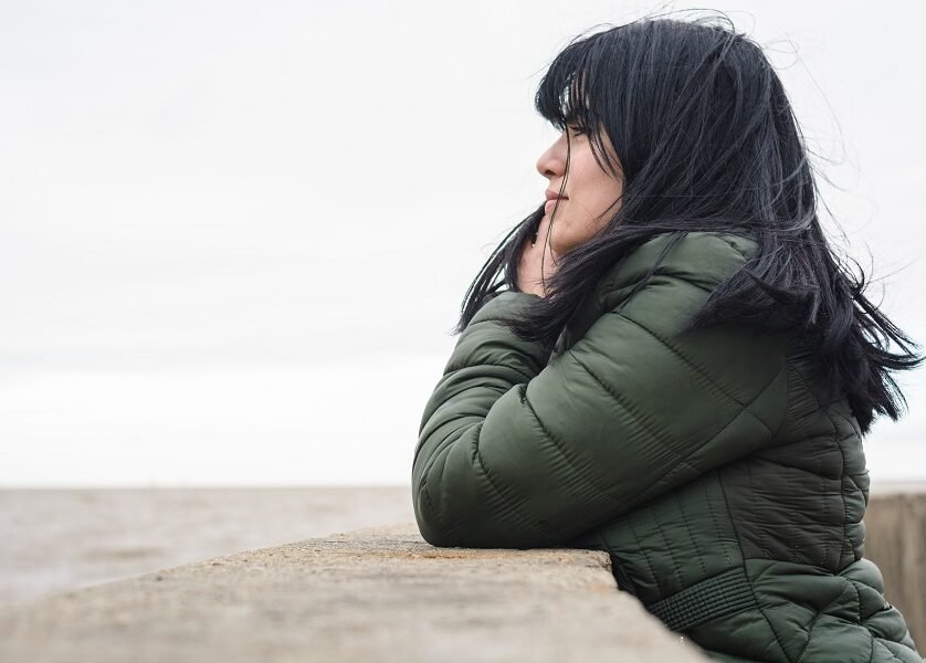 calm young woman in green jacket standing in profile contemplating thinking watching the sea leaning against a wall at the pier on a cloudy afternoon, copy space.